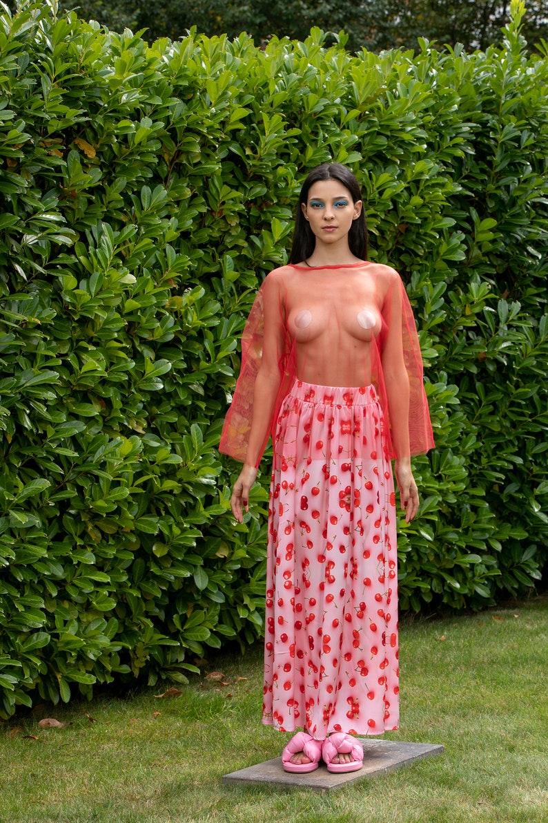 May include: A woman wearing a red sheer top and a pink skirt with a cherry print. She is wearing pink platform sandals.