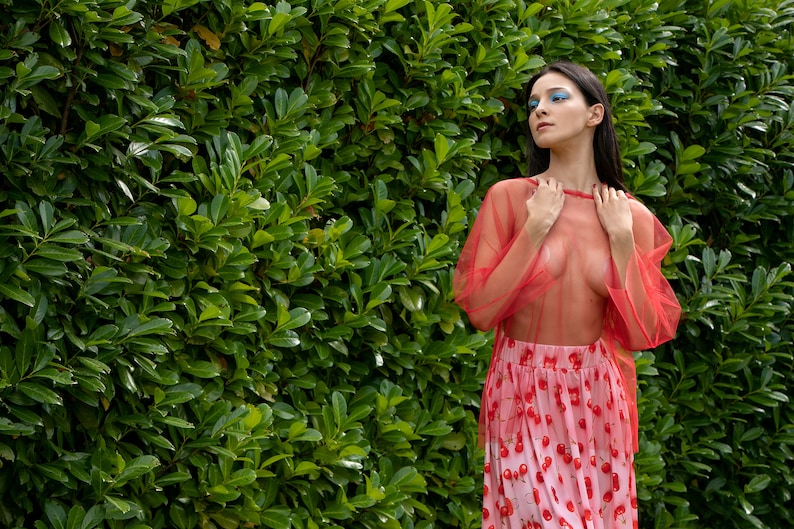 May include: A woman wearing a red sheer top with long sleeves and a cherry print skirt. She is standing in front of a green leafy background.