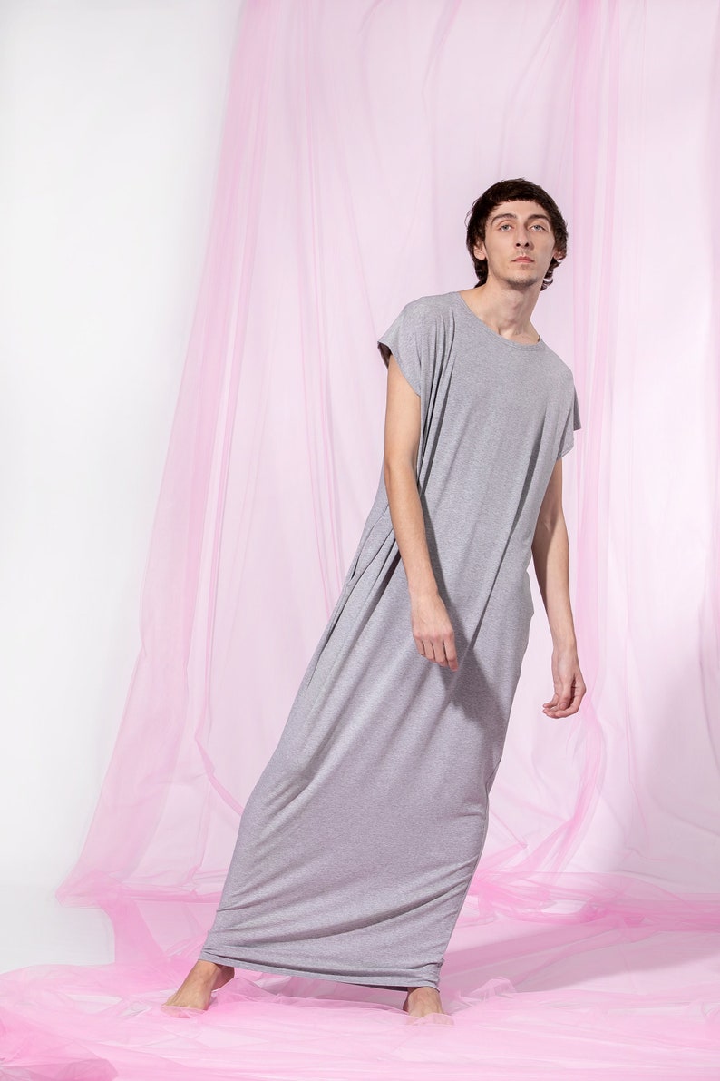 May include: A person wearing a long, gray, loose-fitting dress with short sleeves. The person is standing in front of a pink sheer curtain.