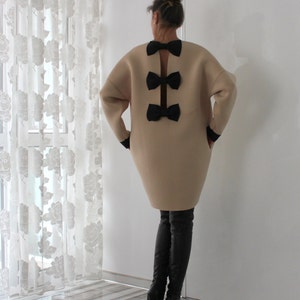 Op de afbeelding: Een beige oversized trui-jurk met een rugdetail van drie zwarte strikken. De jurk wordt gedragen met zwarte laarzen tot aan de dij.