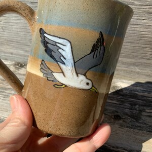 May include: A brown ceramic mug with a white seagull design. The mug has a brown, tan, and blue glaze.