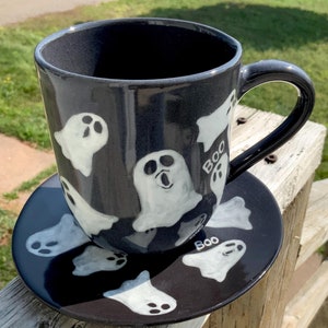 May include: A black ceramic coffee mug and saucer with a Halloween theme. The mug and saucer are decorated with white ghost silhouettes and the word "BOO" written on the mug and saucer.