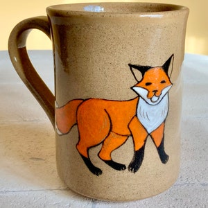 May include: A brown ceramic mug with a hand-painted orange fox design. The fox has black eyes, a white chest, and black legs.