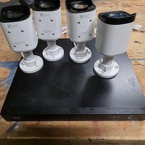 May include: A black rectangular digital video recorder base unit with four white bullet-style security cameras positioned on top. Each camera features a black lens housing, a mounting base, and printed QR codes with technical specifications.