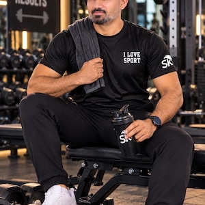 Könnte beinhalten: Ein schwarzes, kurzärmeliges Sport-T-Shirt mit dem weißen Schriftzug I LOVE SPORT auf der Brust und SK auf dem Ärmel. Das Shirt wird mit einer schwarzen Jogginghose und weißen Turnschuhen kombiniert, während ein dunkelgraues Handtuch auf der Schulter liegt.