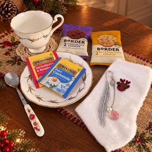 May include: A white porcelain teacup and saucer with gold trim, a silver spoon with a floral handle, two Twinings tea bags, two Border biscuit packets, and a white fabric stocking featuring a metal feather bookmark and a small reindeer patch.