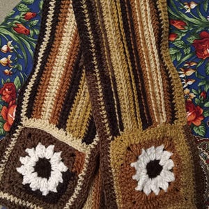 May include: A long, rectangular crochet scarf featuring a vertical striped pattern in shades of dark brown, tan, and cream. Each end of the scarf is finished with a square granny square motif displaying a white flower with a dark brown center.