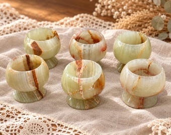 May include: A set of six small, rounded cups crafted from light green stone with prominent brown and white veining. Each cup features a smooth, polished finish, a wide circular opening, and a sturdy, integrated pedestal base for stability.