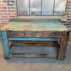 Puede incluir: Un banco de trabajo de madera rústica con tres cajones. La superficie superior está pintada en tonos azules y verdes, con desgaste visible. Las patas y el marco son de madera envejecida, lo que añade un toque vintage. El banco de trabajo es un mueble funcional.