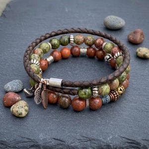 May include: A multi-strand bracelet featuring a dark brown braided leather cord and round beads in shades of green, brown, and orange. The bracelet includes silver-toned metal spacers and two small copper-colored feather charms.