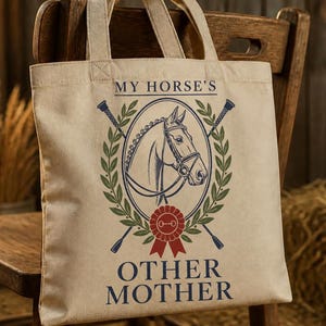 May include: A beige canvas tote bag featuring a graphic of a horse head inside an oval frame, flanked by two crossed riding crops. Below the horse is a red rosette ribbon. The text reads MY HORSE'S OTHER MOTHER in dark blue lettering.
