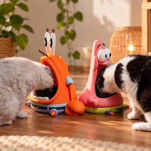 Puede incluir: Dos estaciones de alimentación para gatos con forma de personajes de dibujos animados. Una es un cangrejo naranja con ojos grandes y pinzas, y la otra es una estrella de mar rosa con ojos grandes. Cada estación contiene un cuenco negro.