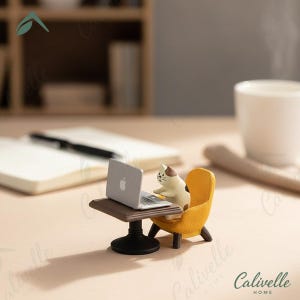 May include: A miniature figurine of a calico cat sitting in a yellow armchair at a small brown table with a silver laptop. The laptop features a small white apple logo on the lid. The cat has brown and white fur markings and is facing the screen.