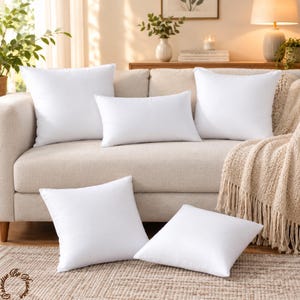 May include: A collection of five plain white scatter cushions in various shapes, including three square cushions and two rectangular cushions, arranged on and in front of a light-coloured sofa with a textured beige throw blanket draped over the side.