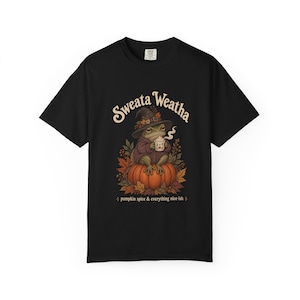 Puede incluir: Una camiseta negra con el gráfico de una rana que lleva un sombrero de bruja y un suéter, sentada sobre una calabaza mientras sostiene una taza humeante. El texto dice Sweata Weatha y pumpkin spice & everything nice-ish en color crema.