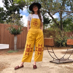 May include: A mustard yellow jumpsuit worn over a white t-shirt. The jumpsuit features a square bib with a brown geometric star pattern and matching star patches on the lower legs. It includes thin shoulder straps and wide-leg trousers.