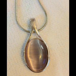 Puede incluir: Un collar con colgante de tono plateado que presenta una piedra cabujón ovalada de color morado claro con un efecto ojo de gato chatoyante. La piedra está engastada en un marco de metal liso conectado a una cadena de serpiente plateada y flexible.