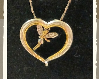 May include: A two-tone gold and silver heart-shaped pendant necklace featuring a small fairy figure suspended in the centre. The fairy has delicate wings adorned with tiny clear stones and is positioned within the open frame of the heart.