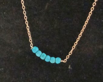 May include: A delicate gold-toned chain necklace featuring a horizontal row of six small, round, bright turquoise-coloured beads centred along the chain. The necklace has a simple link design and the beads are arranged in a slight curve.
