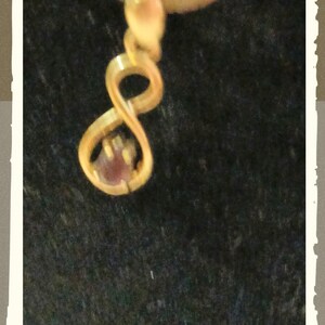 May include: A gold-toned pendant featuring an infinity symbol shape with a small, round, reddish-brown gemstone set within the lower loop of the design. The pendant is suspended from a thin gold-toned chain, creating a delicate jewelry piece.