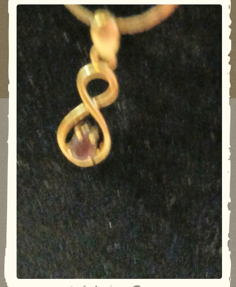 May include: A gold-toned pendant featuring an infinity symbol shape with a small, round, reddish-brown gemstone set within the lower loop of the design. The pendant is suspended from a thin gold-toned chain, creating a delicate jewellery piece.