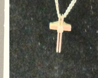 May include: A light-coloured cross pendant suspended from a matching twisted cord necklace. The cross features a vertical bar that extends below the horizontal crossbar, creating a distinct elongated shape. The entire piece is a uniform pale off-white.