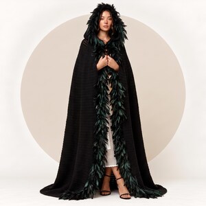 May include: A floor-length black hooded cloak featuring a textured knit fabric and a thick trim of dark green feathers lining the hood and the front opening. The garment drapes to the floor and includes a deep hood with matching feather accents.