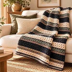 May include: A crocheted throw blanket featuring horizontal stripes in alternating colors of cream, dark charcoal, sage green, and light brown. The blanket has a textured, open-work stitch pattern and is draped over the seat of a light-colored sofa.