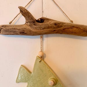 May include: A decorative wall hanging featuring a driftwood branch with a light green ceramic fish pendant. The fish has a textured scale pattern and a wooden bead accent. The piece is suspended by twine.