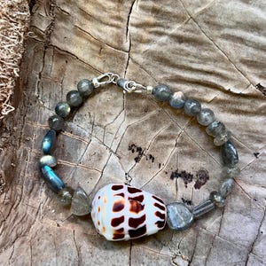 May include: A handmade bracelet featuring a white and brown seashell centerpiece. The bracelet is strung with gray and blue-green gemstone beads and has a silver clasp. The bracelet is displayed on a textured, wooden surface.