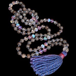 Könnte beinhalten: Eine Perlenkette mit abwechselnden runden, gesprenkelten braunen Steinperlen und durchscheinenden, irisierenden Glasperlen. Die Kette endet in einer großen, dicken Quaste aus blauem und violettem Garn, die mit einem dünnen roten Faden befestigt ist.