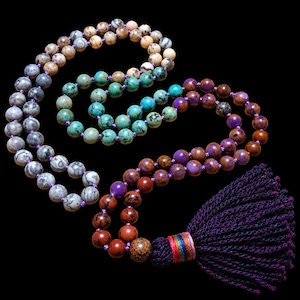 Könnte beinhalten: Eine lange Mala-Kette mit verschiedenen runden Steinperlen in Grau-, Beige-, Türkis-, Violett- und Rotbrauntönen. Die Perlen sind auf eine violette Schnur gefädelt und enden in einer großen, dunkelvioletten Quaste mit einer bunten Wicklung.