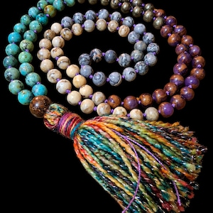 Könnte beinhalten: Eine lange Perlenkette mit verschiedenen runden Steinen in den Farben Türkis, Metallic-Gold, Grau, Beige und Violett. Die Perlen sind auf eine violette Schnur gefädelt und mit einer großen, bunten Quaste aus gedrehtem Garn versehen.