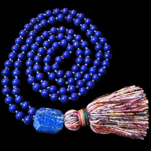 Könnte beinhalten: Eine lange Halskette mit zahlreichen runden, tiefblauen Lapislazuli-Perlen, akzentuiert durch einen großen, facettierten hexagonalen Lapislazuli-Anhänger und eine dicke, mehrfarbige Quaste aus weißen, rosafarbenen und blauen Fäden.