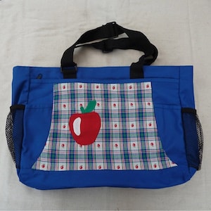 May include: A blue tote bag with a plaid pocket featuring a red apple applique. The bag has two black straps and mesh side pockets.