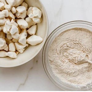 May include: A light-coloured bowl filled with irregular, off-white chunks of baobab fruit pulp next to a clear glass bowl containing a fine, beige powder mixture with a metal spoon resting inside, showcasing the raw and processed fruit components.