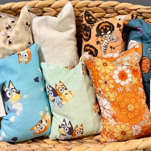 Peut inclure: Un ensemble de sept sacs de riz en tissu rectangulaires avec divers motifs incluant des chiens de dessin animé, des fleurs orange et des thèmes d'Halloween, présentés dans un panier tressé avec deux cartes informatives Create with Kate et Rice Bag.