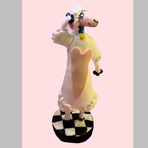 May include: A white sheep figurine with a peach-colored heart on its chest, wearing a blue collar with a yellow tag. The sheep has black hooves, a pink face, and is standing on a circular base with a black and white checkered pattern.