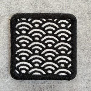 May include: A square embroidered patch featuring a repeating black and white pattern of stylized concentric semicircles. The design is bordered by a thick, black stitched edge, creating a defined frame around the geometric wave-like motif.