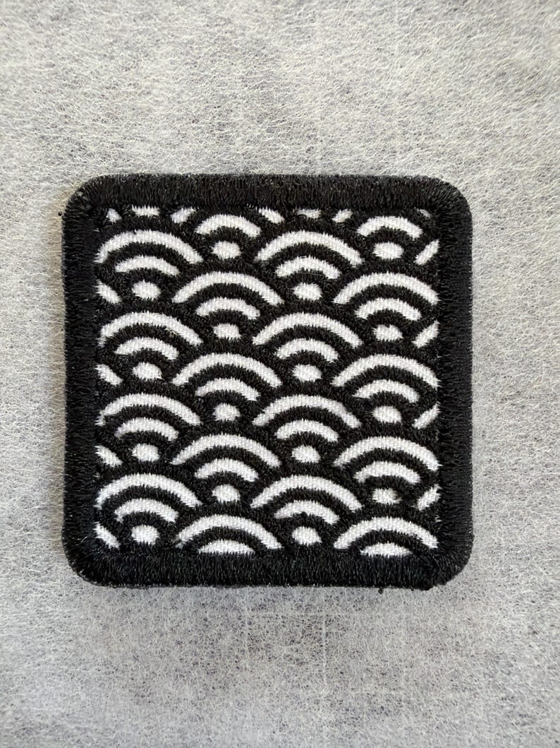 May include: A square embroidered patch featuring a repeating black and white pattern of stylized concentric semicircles. The design is bordered by a thick, black stitched edge, creating a defined frame around the geometric wave-like motif.