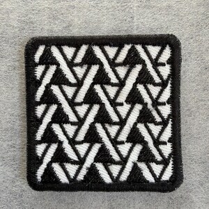 May include: A square embroidered patch featuring a repeating geometric pattern of interlocking black and white lines. The design consists of angled segments that create a series of triangles and diagonal bars, all bordered by a thick black stitched edge.