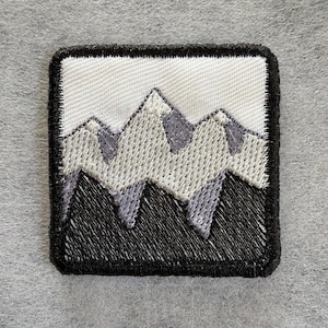 May include: A square embroidered patch featuring a mountain range design. The patch has a thick black border, a white sky background, light gray snow-capped peaks, and solid black mountain bases, all rendered in detailed thread embroidery.