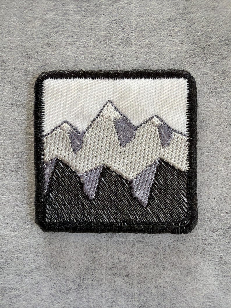 May include: A square embroidered patch featuring a mountain range design. The patch has a thick black border, a white sky background, light gray snow-capped peaks, and solid black mountain bases, all rendered in detailed thread embroidery.