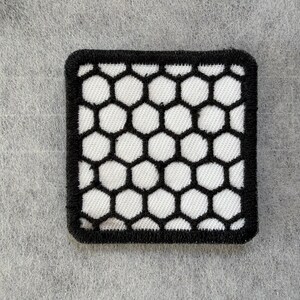 May include: A square embroidered patch featuring a repeating black honeycomb pattern over a white background. The patch has a thick, solid black stitched border that frames the geometric hexagonal design, creating a high-contrast visual effect.