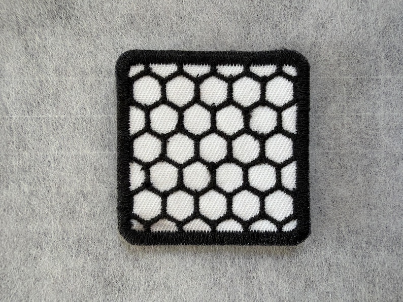 May include: A square embroidered patch featuring a repeating black honeycomb pattern over a white background. The patch has a thick, solid black stitched border that frames the geometric hexagonal design, creating a high-contrast visual effect.