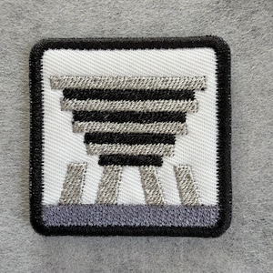 May include: A square embroidered patch featuring a white background with a black border. The design depicts a stylised, inverted pyramid shape composed of alternating horizontal black and silver-grey stripes, positioned above three silver-grey vertical bars.