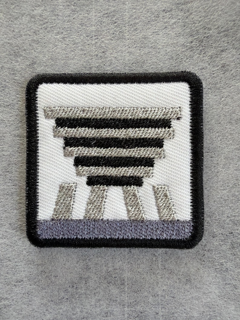Puede incluir: Un parche bordado cuadrado con fondo blanco y borde negro. El dise&ntilde;o muestra una forma de pir&aacute;mide invertida estilizada compuesta por rayas horizontales alternas negras y gris plateado, situada sobre tres barras verticales de color gris plateado.