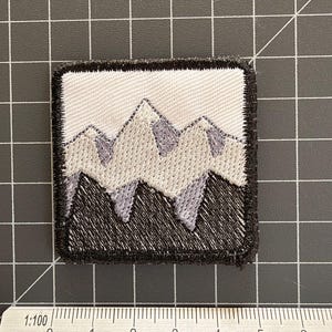 May include: A square embroidered patch featuring a mountain range design in shades of white, light gray, and black. The patch is positioned above a ruler showing a width of approximately 4.5 centimeters. The design has a thick black border.