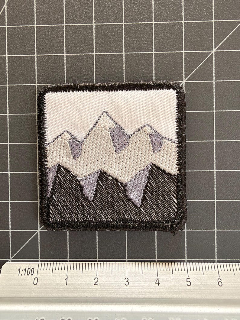 May include: A square embroidered patch featuring a mountain range design in shades of white, light gray, and black. The patch is positioned above a ruler showing a width of approximately 4.5 centimeters. The design has a thick black border.