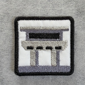May include: A square embroidered patch featuring a white background with a black border. The design depicts a stylised grey and black structure resembling a torii gate or architectural element with horizontal bars and vertical supports.